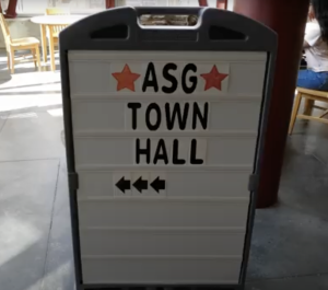A standing sign with a grey frame displays black text reading "ASG TOWN HALL" on a white background. Below the text, three black left-pointing arrows indicate direction a College of the Canyons